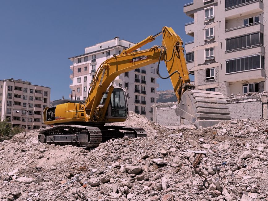 Yellow excavator working on urban construction site amidst rubble and buildings.
