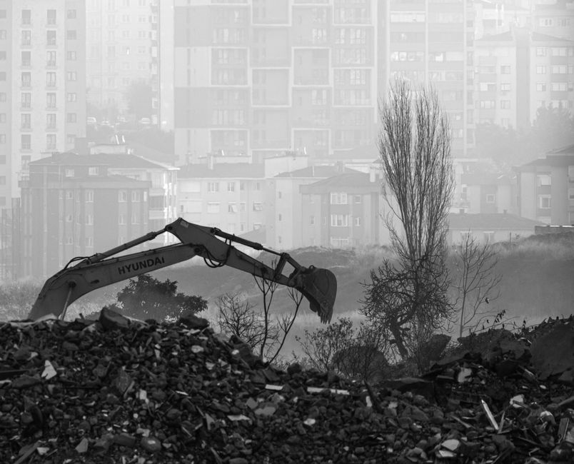 An excavator working on a cleared site with buildings in the distance.